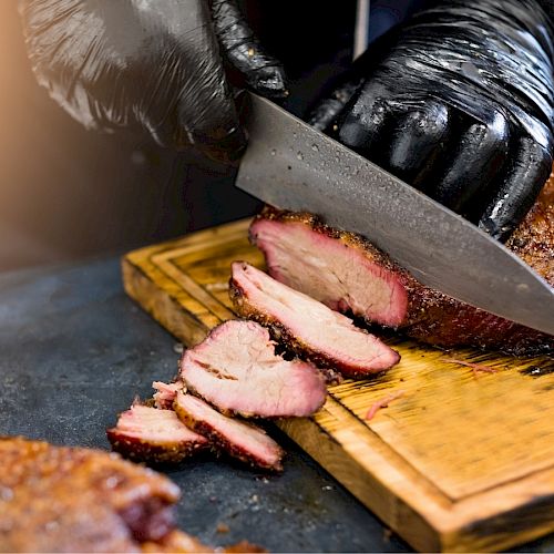 A person wearing black gloves is slicing smoked meat on a wooden cutting board with a knife.