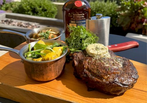 A grilled steak with herb butter, accompanied by vegetables in pots, and a bottle in the background, served on a wooden board.