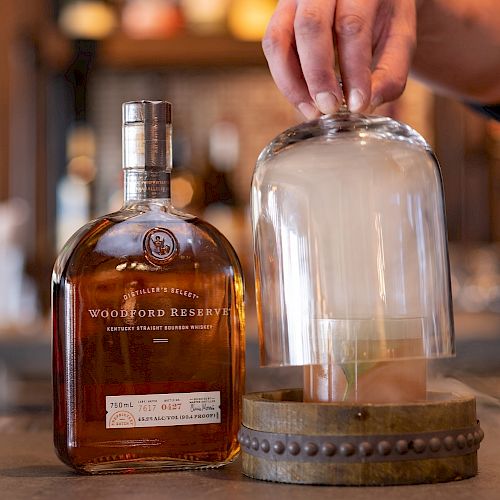 A bottle of bourbon and a cocktail under a glass dome with smoke, beside a hand lifting the dome in a bar setting.