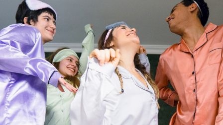 Four people in colorful pajamas are joyfully dancing together, each wearing a sleep mask on their heads, in a fun indoor setting.