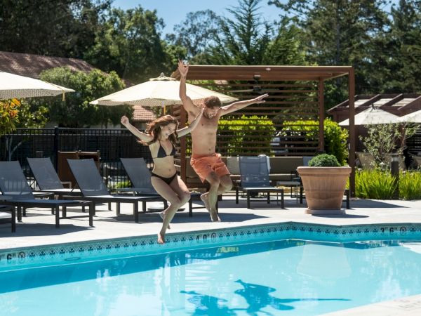 Two people are jumping into a pool from the edge, mid-air, with sun loungers, umbrellas, and a sunny resort vibe in the background.