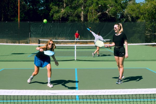 People are playing pickleball on an outdoor court, with a player about to hit the ball, while others watch and participate in the game.