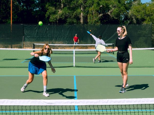 People are playing pickleball on an outdoor court, with a player about to hit the ball, while others watch and participate in the game.