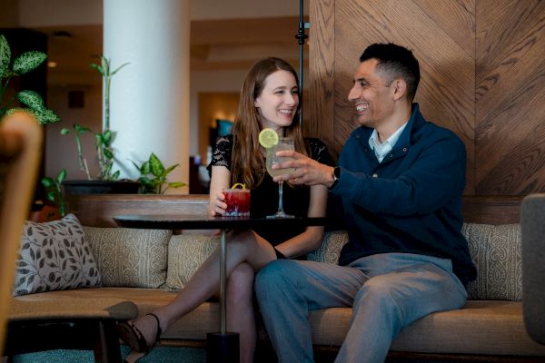 A couple is sitting on a sofa, smiling and holding drinks. The setting is cozy with plants and a wooden backdrop.