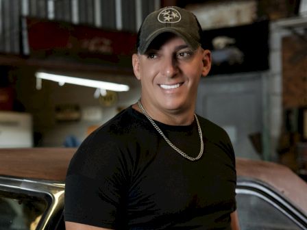 A person in a black shirt and cap smiles, standing next to a car in a garage.