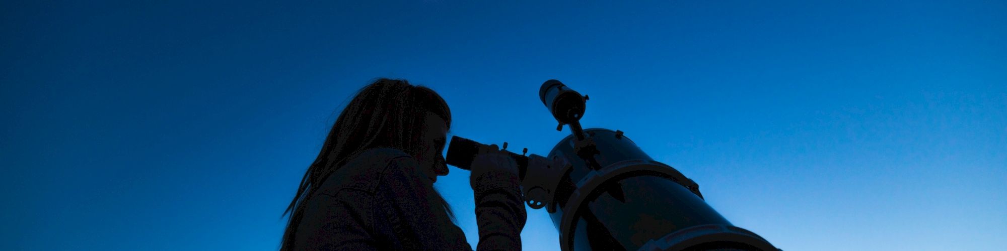 A person is using a telescope to observe the crescent moon in a clear night sky.
