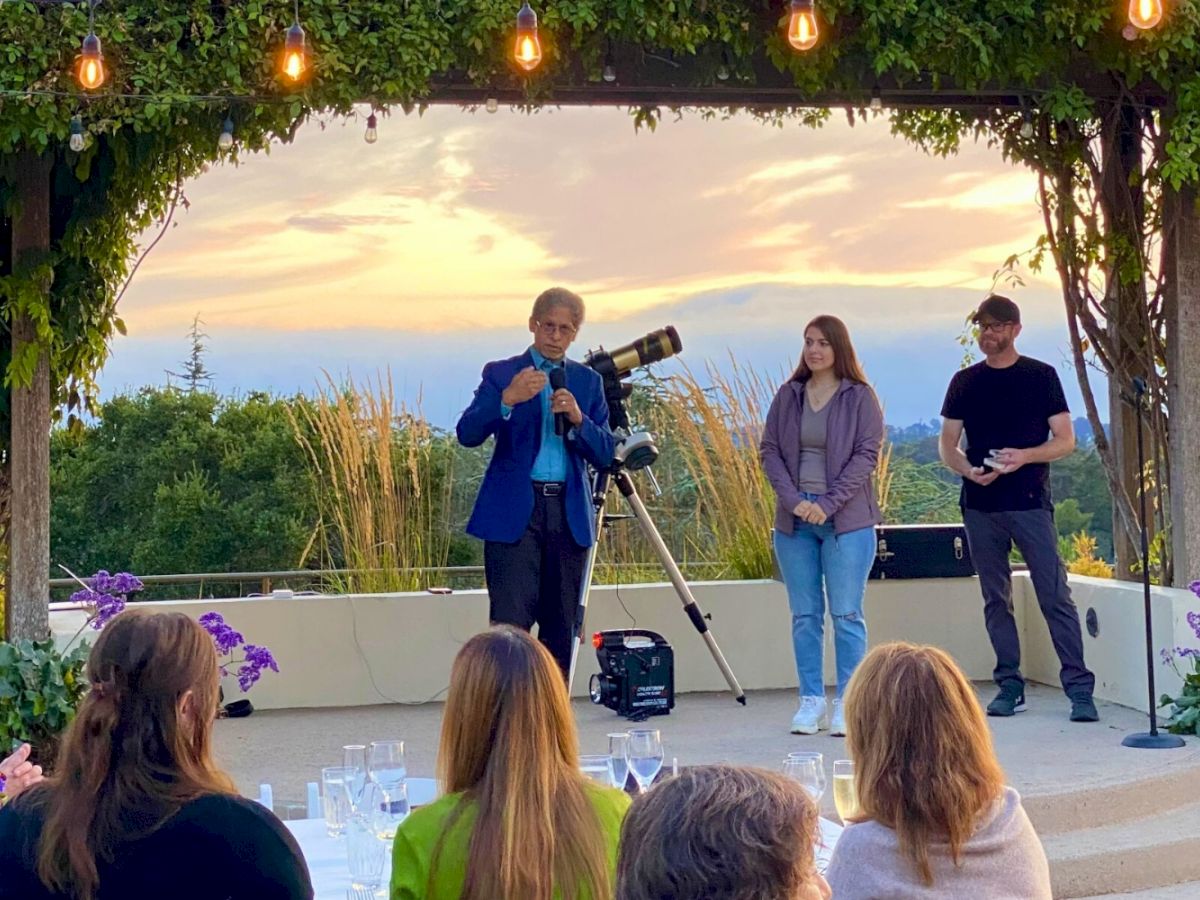 A group of people are gathered outdoors at sunset, listening to a speaker with a telescope nearby.