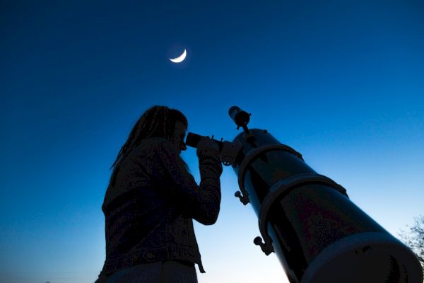 A person is using a telescope to gaze at the crescent moon against a clear, twilight sky.
