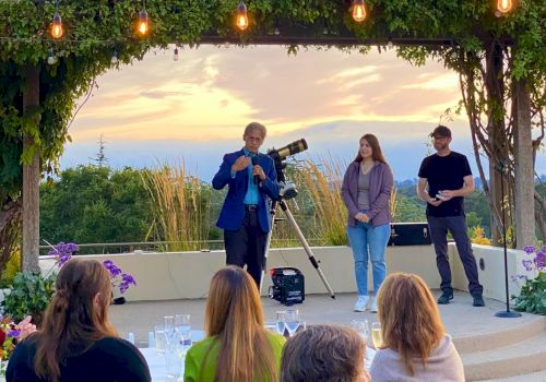 A person speaks to an audience outdoors, with two others nearby and a telescope in the background under a vine-covered pergola.