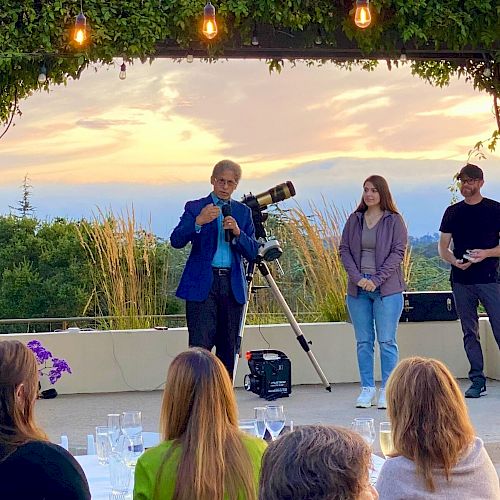 A person speaks to an audience outdoors, with two others nearby and a telescope in the background under a vine-covered pergola.
