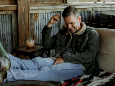 Man relaxing on a cushioned seat with a drink beside, wearing a jacket and jeans, smiling and looking comfortable in a rustic setting.