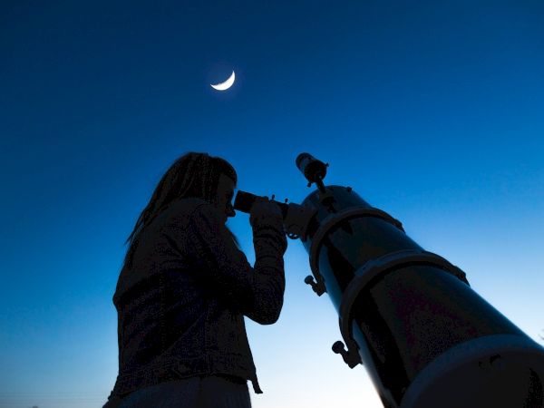 A person is using a telescope to observe the crescent moon in the evening sky, silhouetted against the twilight.