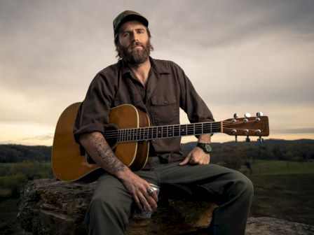 A person with a beard sits outdoors on a rock, holding an acoustic guitar and a can, under a cloudy sky.