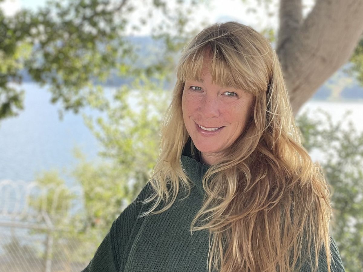 A person with long hair stands outdoors by a lake, surrounded by trees. The person wears a green sweater and smiles at the camera.