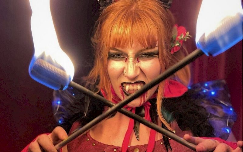 A person with fiery red hair is holding crossed flaming torches, wearing elaborate makeup and attire, looking intensely at the viewer.