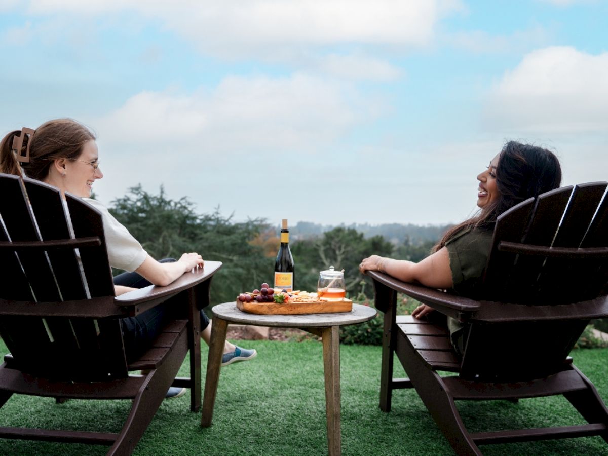 Two people sitting in outdoor chairs, with food and wine on a table, enjoying a scenic view.