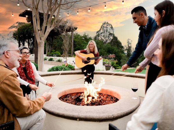 A group of people are enjoying a fire pit outdoors, with one playing guitar, others roasting marshmallows, against a scenic sunset backdrop.