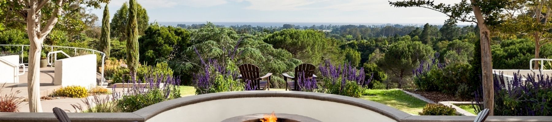 A scenic patio with a circular fire pit, surrounded by four chairs, overlooks a lush green landscape under a partly cloudy sky.