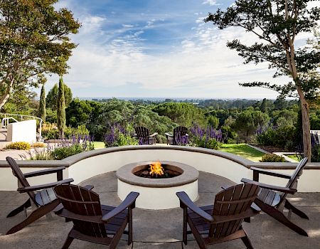 A scenic patio with a circular fire pit, surrounded by four chairs, overlooks a lush green landscape under a partly cloudy sky.