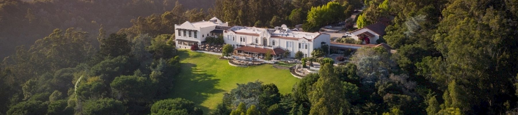 A lavish estate with white buildings sits on a grassy lawn surrounded by dense forest and rolling hills, basking in clear blue sky.