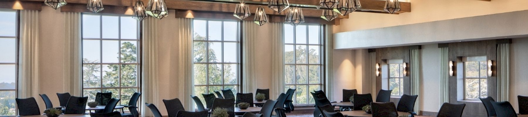 A bright conference room with round tables, black chairs, hanging pendant lights, carpet, and large windows&mdash;ready for meetings and events.