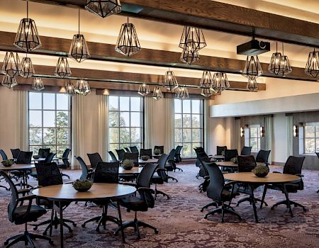 A bright conference room with round tables, black chairs, hanging pendant lights, carpet, and large windows&mdash;ready for meetings and events.