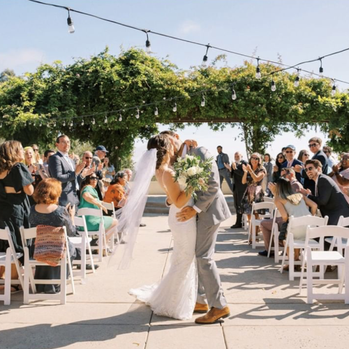 A couple in wedding attire kiss under a greenery arch as guests sit on white chairs and cheer, outdoors on a sunny day with string lights.