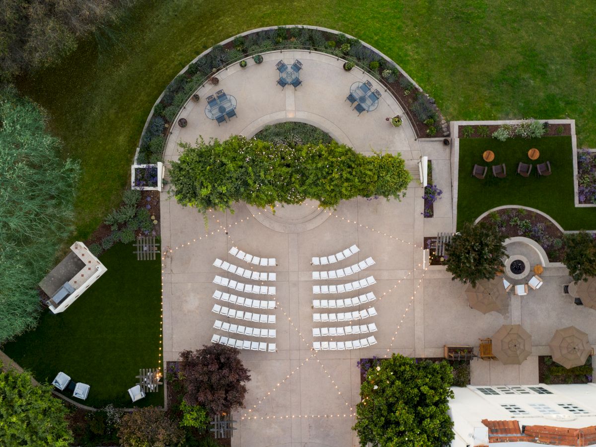 An aerial view shows an outdoor event setup: rows of white chairs on a courtyard, curved greenery, a circular seating area, and surrounding lawn and buildings.