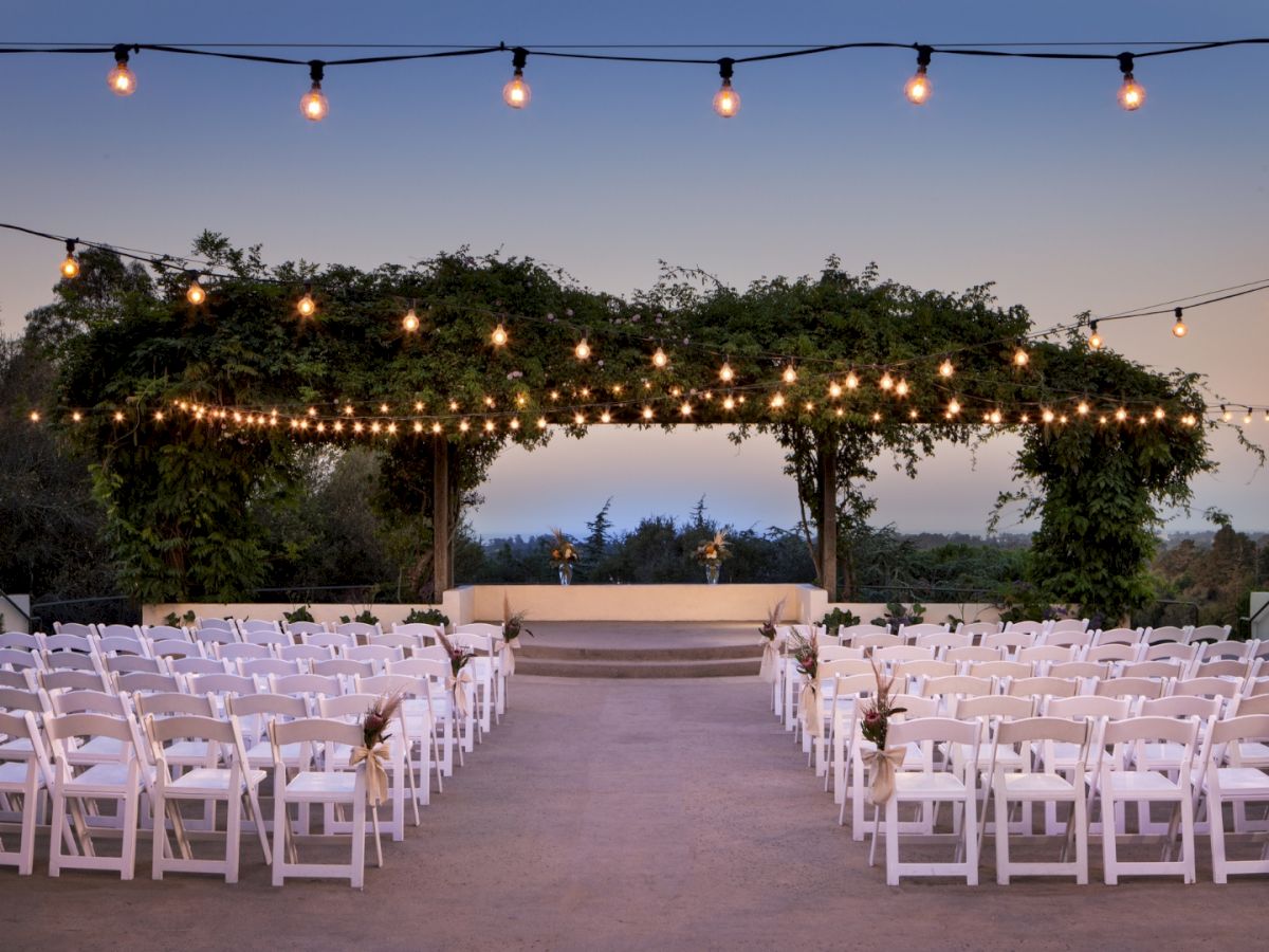 Outdoor wedding setup with string lights, a floral arbor, and white chairs arranged in rows facing a stage, serene horizon in background.