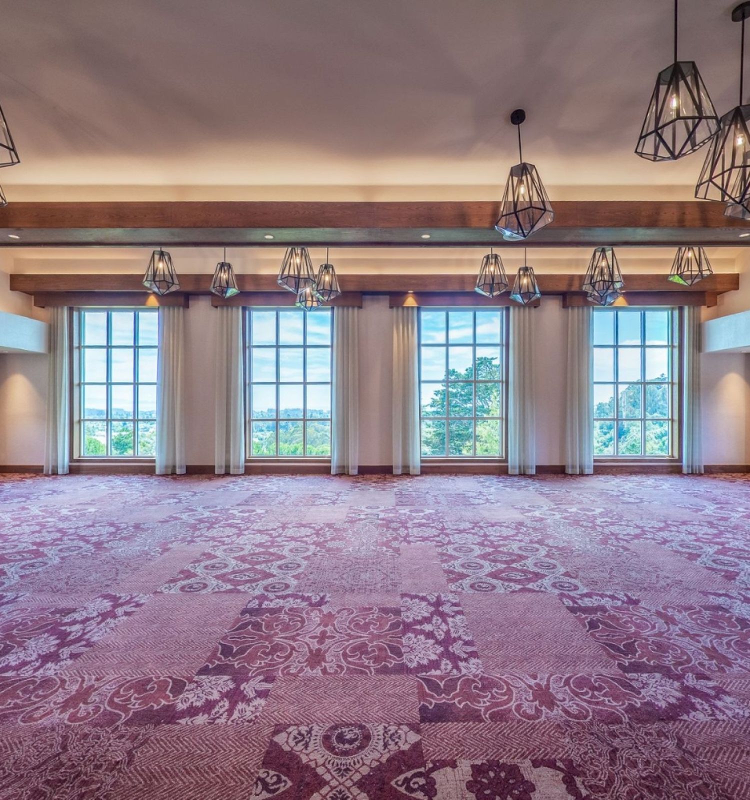 A large empty banquet hall with floral carpet, hanging geometric chandeliers, wooden beams, and large windows letting in natural light.