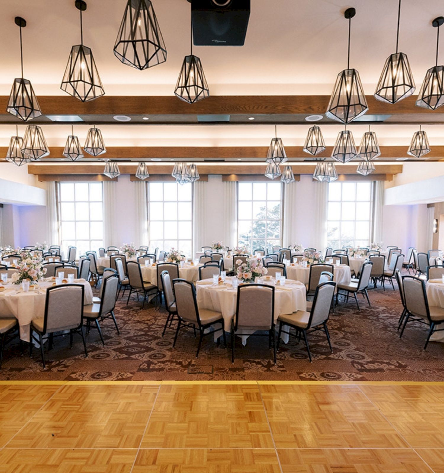 A spacious banquet hall set for a formal event, with round tables, beige chairs, warm lighting, and a polished wooden stage area in the foreground.