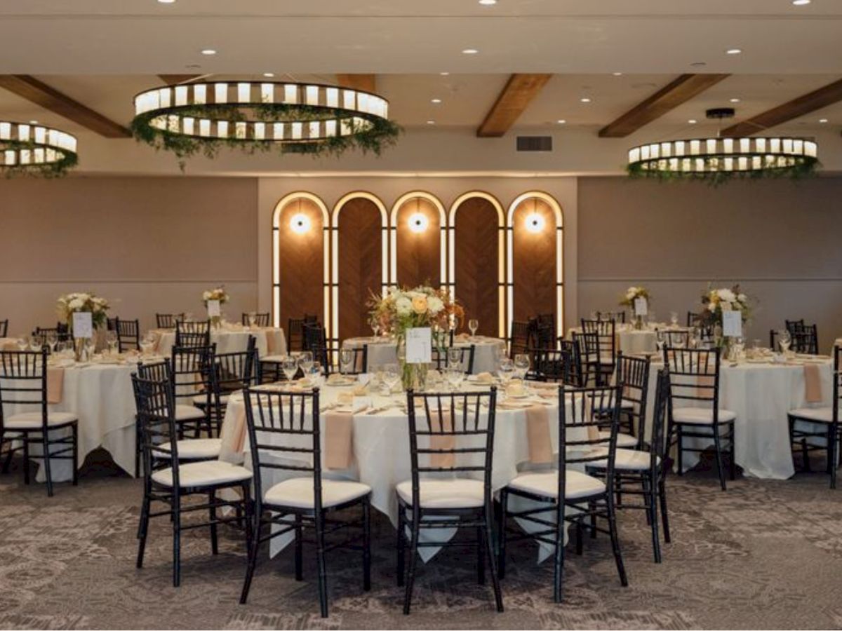 A stylish banquet hall with round tables, black chairs, white tablecloths, floral centerpieces, and warm lighting in an elegant setting.