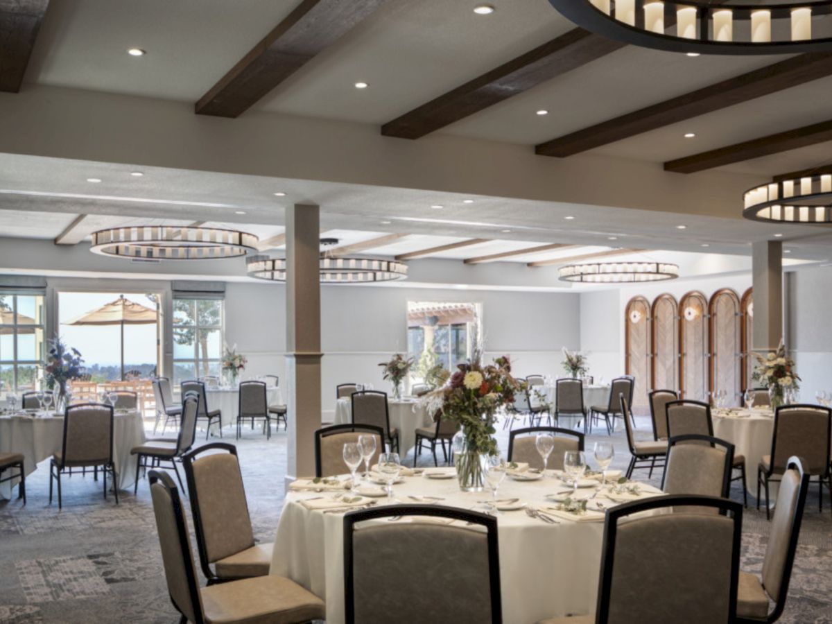 A elegant banquet hall with round tables, cream chairs, floral centerpieces, and warm lighting under wooden ceiling beams, ready for a formal event.