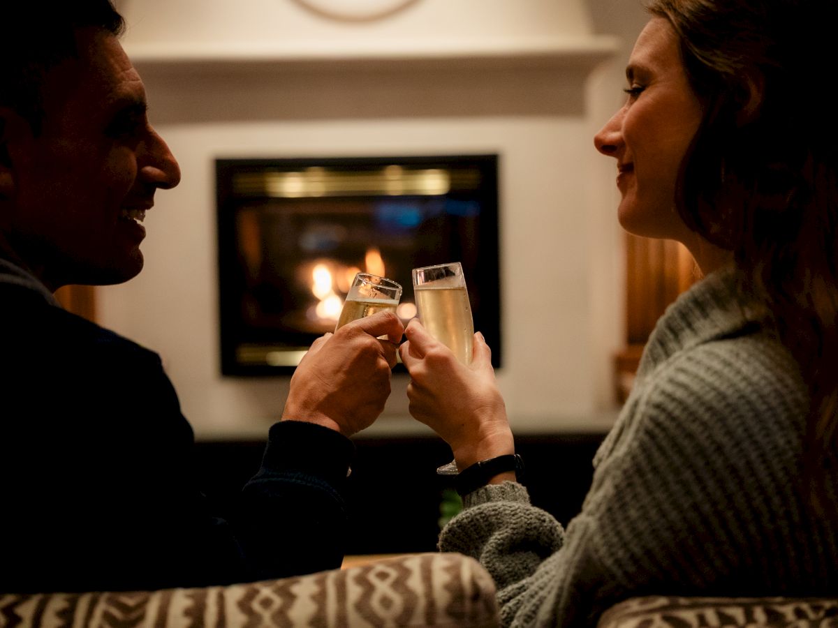 Two people clink champagne flutes by a lit fireplace, sharing a cozy, intimate moment on a sofa.