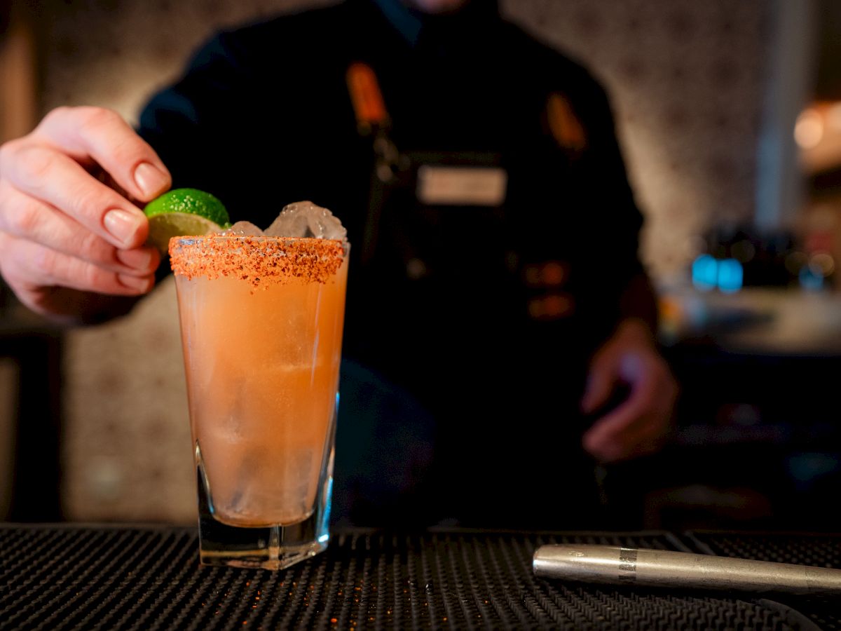 A bartender adding a lime wedge to a frosty peach cocktail with a salted rim, on a bar counter, metal tool nearby, dim background.