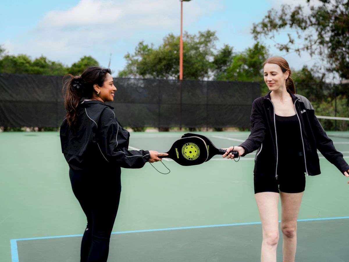 Two friends on a tennis court share a high-five with a retractable tethered ball spinner between them, smiling after a playful moment.