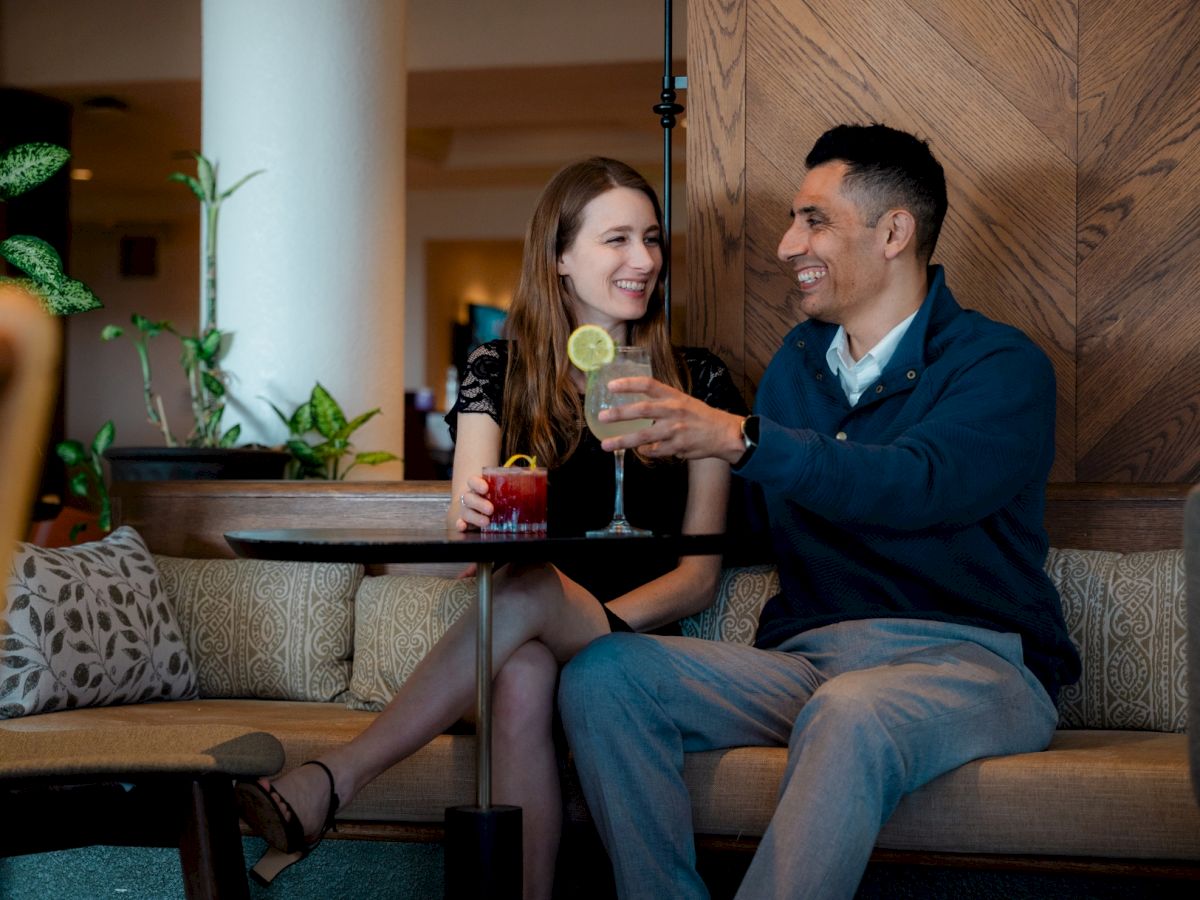 Two people are enjoying drinks together on a sofa at a bar, smiling and chatting in a relaxed lounge setting.