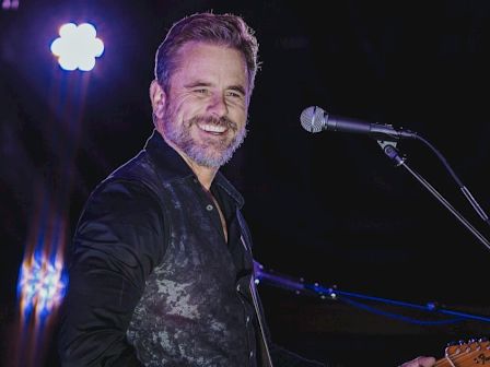 A smiling male musician on stage with a guitar, wearing a dark vest, microphone stand nearby, spotlighted backdrop.