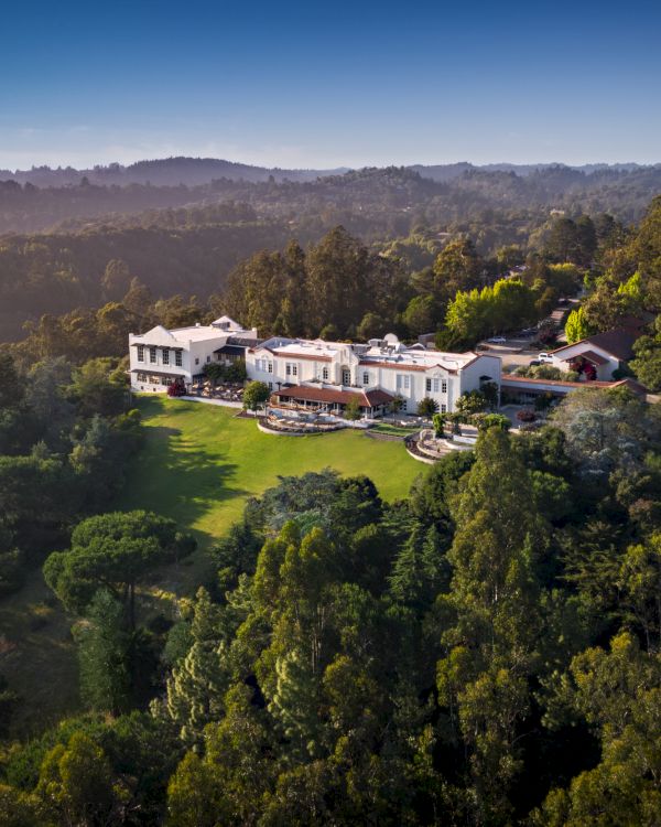 Luxurious mansion complex nestled in dense forest, white buildings with manicured lawn surrounded by tall trees and distant hills.