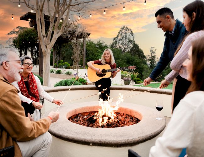 Group of diverse friends sits around a fire pit while a guitarist plays at sunset, with string lights overhead.