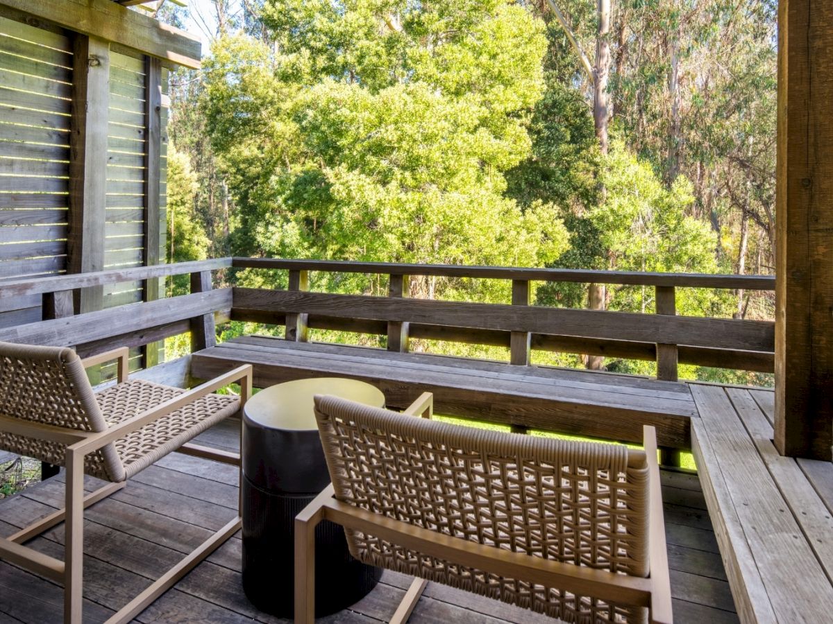 A wooden balcony with woven chairs around a small round table, overlooking green trees and sunny scenery outside.