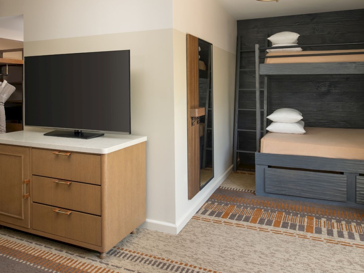 Hotel room with a dresser and TV on the left, a full-length mirror, and a dark bunk with a mattress and pillows on the right.