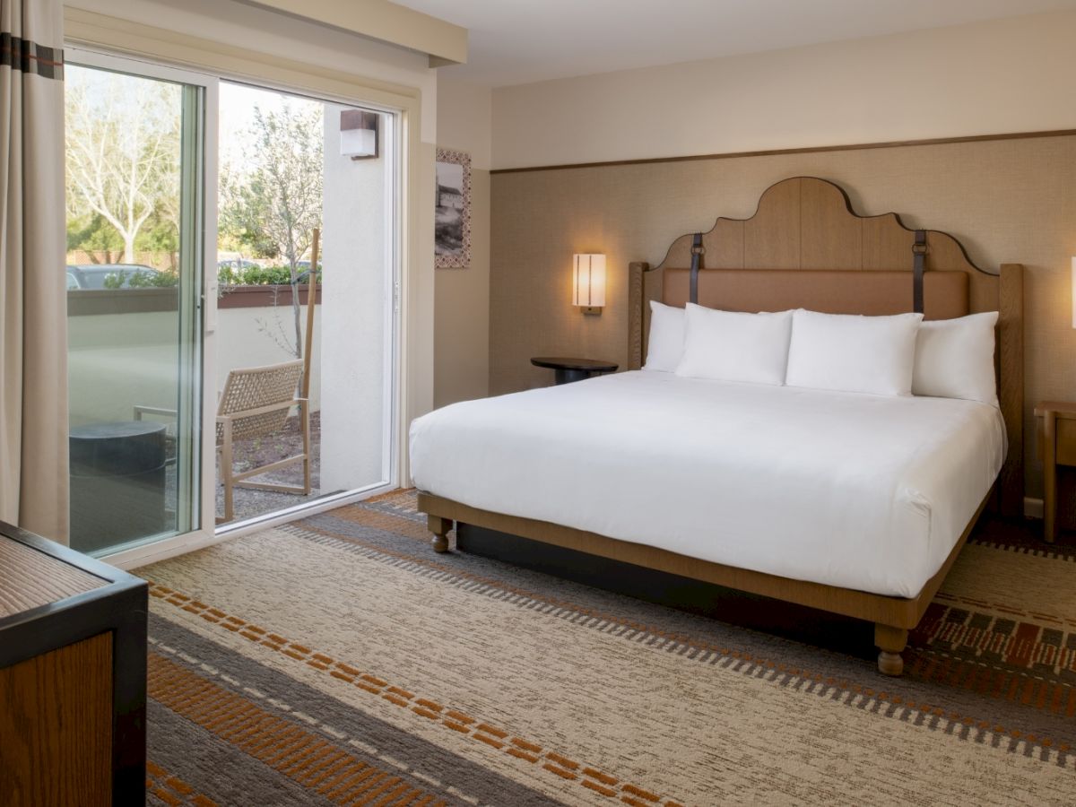 A hotel room with a large bed, wooden headboard, nightstands, lamps, and sliding glass door to a balcony, overlooking trees.
