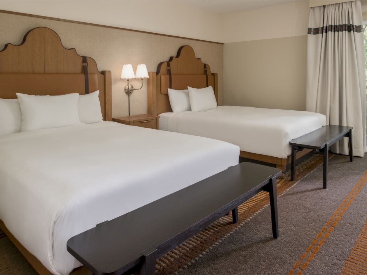 Two large beds with white bedding and wooden headboards, a bench at each foot, bedside lamps, and a window with curtains in a hotel room.