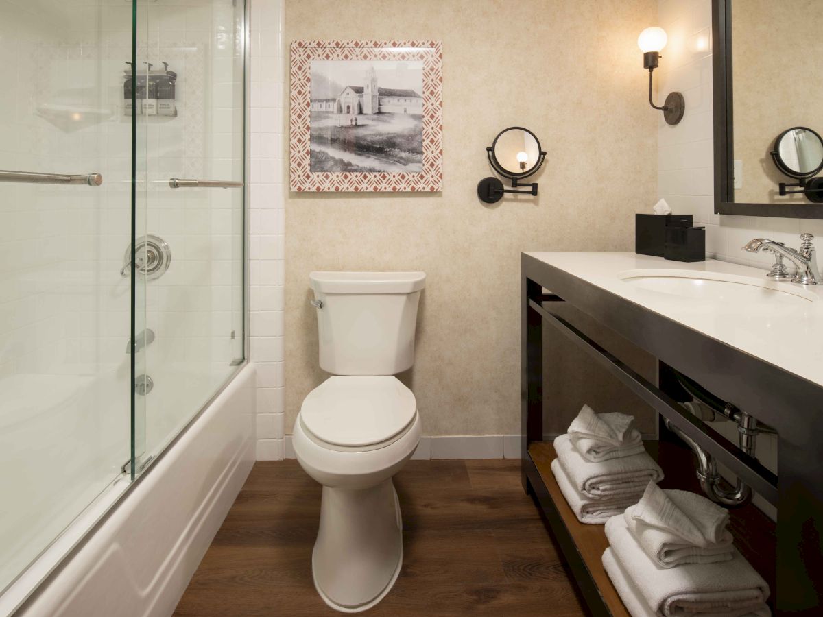 A modern bathroom with a glass shower, a white toilet, and a dark wood vanity with open shelves and towels, plus framed art on the wall.