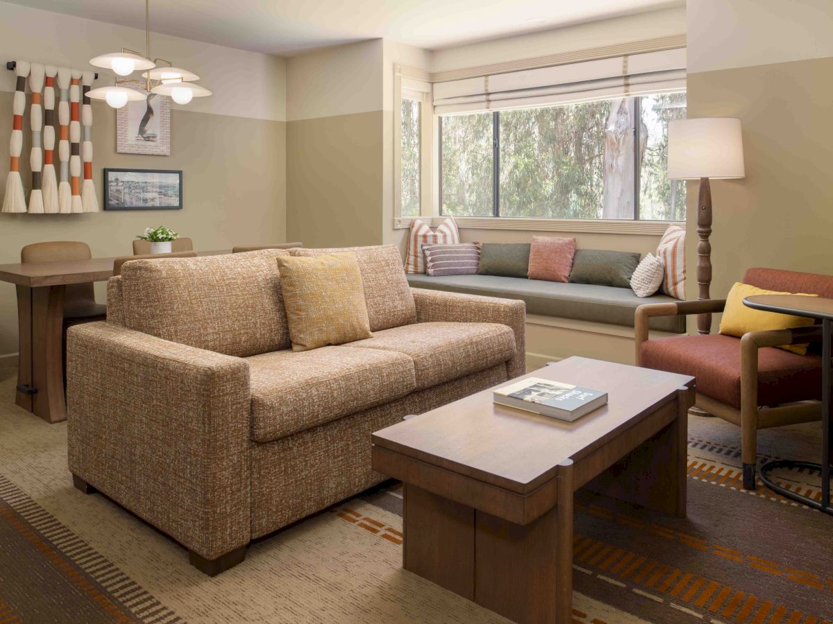 Cozy living room with a beige sofa, matching loveseat, wooden coffee table, and a windowed bench seat; warm lighting and colorful accents.