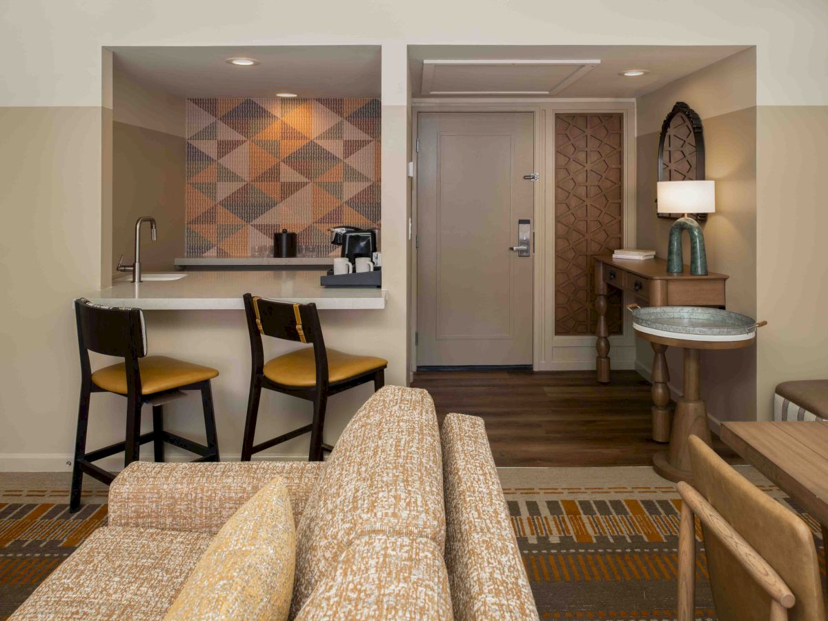A hotel suite living area with a small kitchenette, geometric accent wall, two bar stools at a counter, a beige sofa, and a hallway with a console table and mirror.-ending sentence.