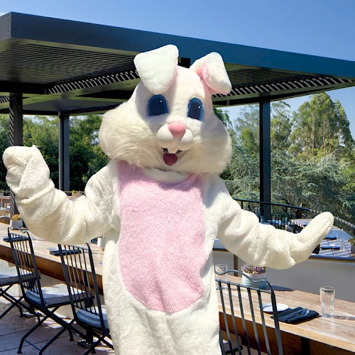 A person in a white and pink bunny costume is posing outside on a patio with tables and chairs.