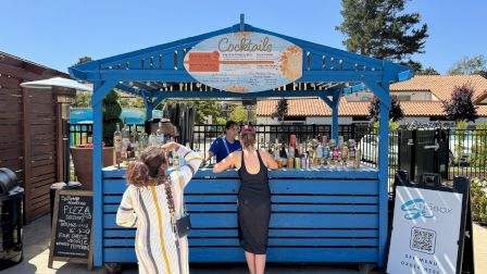 A blue outdoor cocktail stand with two customers ordering drinks in a sunny setting. There's signage and various bottles on display.
