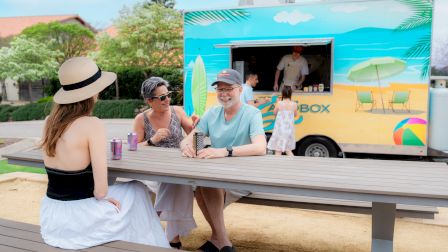 A sunny beachside food truck serves up smiles as friends chat at a picnic table, sun hats on, blue vibes, and casual drinks.
